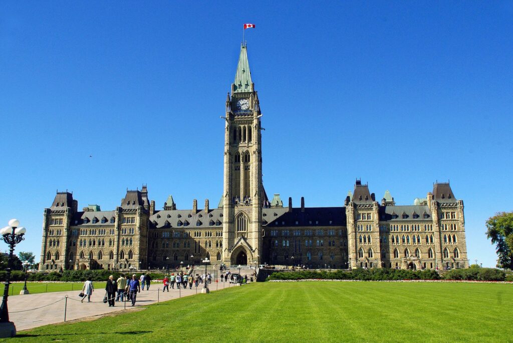 A photo of the Canadian Parliament Buildings in Ottawa, with a clear blue sky, green lawn in the foreground, and people walking on the pathways.