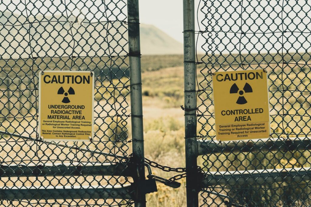 A chain-link fence with caution signs warns of underground radioactive material, with a blurred landscape behind.