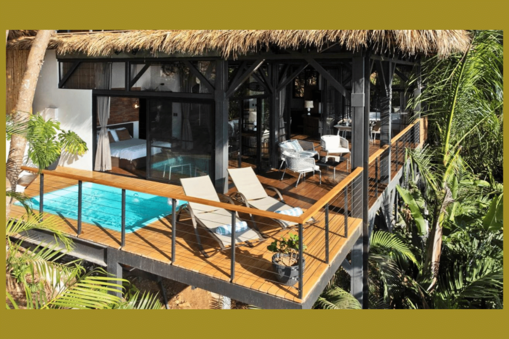 A tropical boutique resort with a thatched roof, featuring a balcony with sun loungers overlooking a private pool, surrounded by lush vegetation.