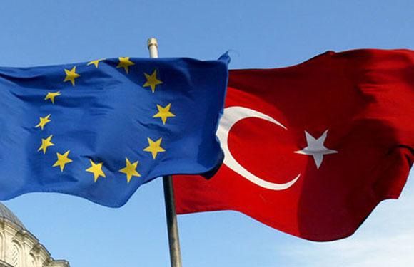 Two flags fly: one blue with yellow stars for the EU, the other red with a white star and crescent for Turkey.