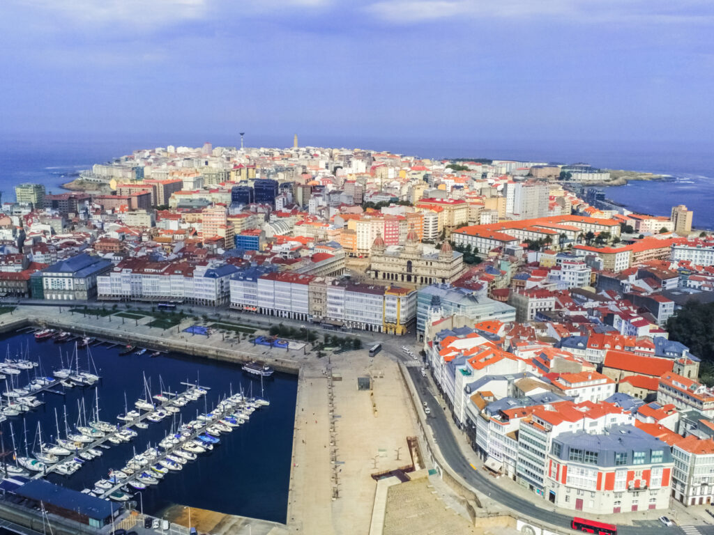 Aerial view of A Coruña, a coastal city in Spain, with dense urban architecture, marina with boats, and clear skies.