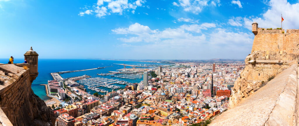 A high vantage point offers a panoramic view of Alicante, Spain, featuring the cityscape, marina, and Mediterranean Sea under a clear sky.