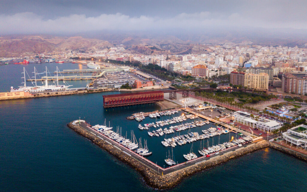 Aerial view of a coastal city, likely Almeria, with a boat-filled marina, urban buildings towards mountains, under a cloudy sky.