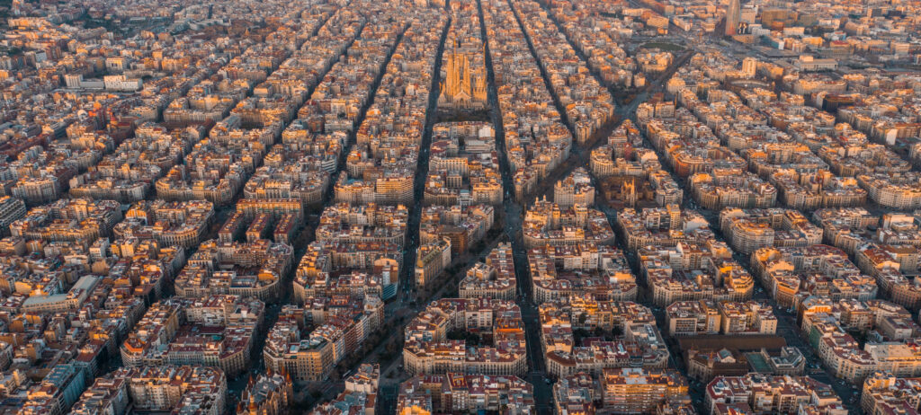 Aerial view of Barcelona's cityscape at dusk, showcasing the grid-like pattern of streets and blocks with warm lighting.