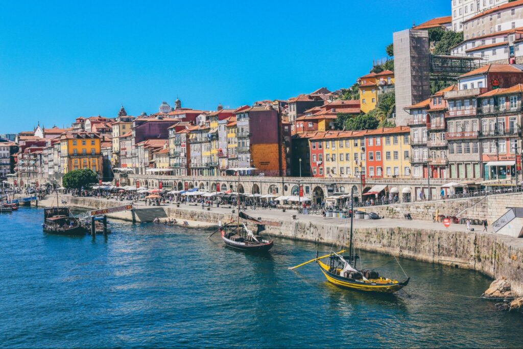 A scenic view of a river with a traditional boat, flanked by colorful buildings on a hillside, likely in Portugal.