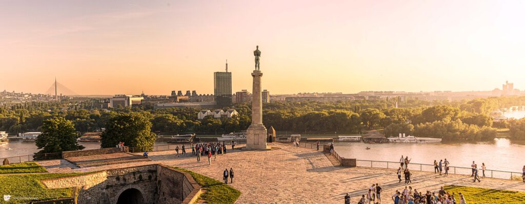 A panoramic view of a cityscape at sunset with a prominent monument and river, likely in Serbia, Europe,.