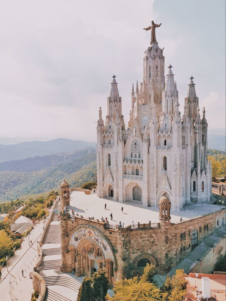 A grand, ornate church with multiple spires and a statue, set against greenery and a clear sky, possibly in Spain.