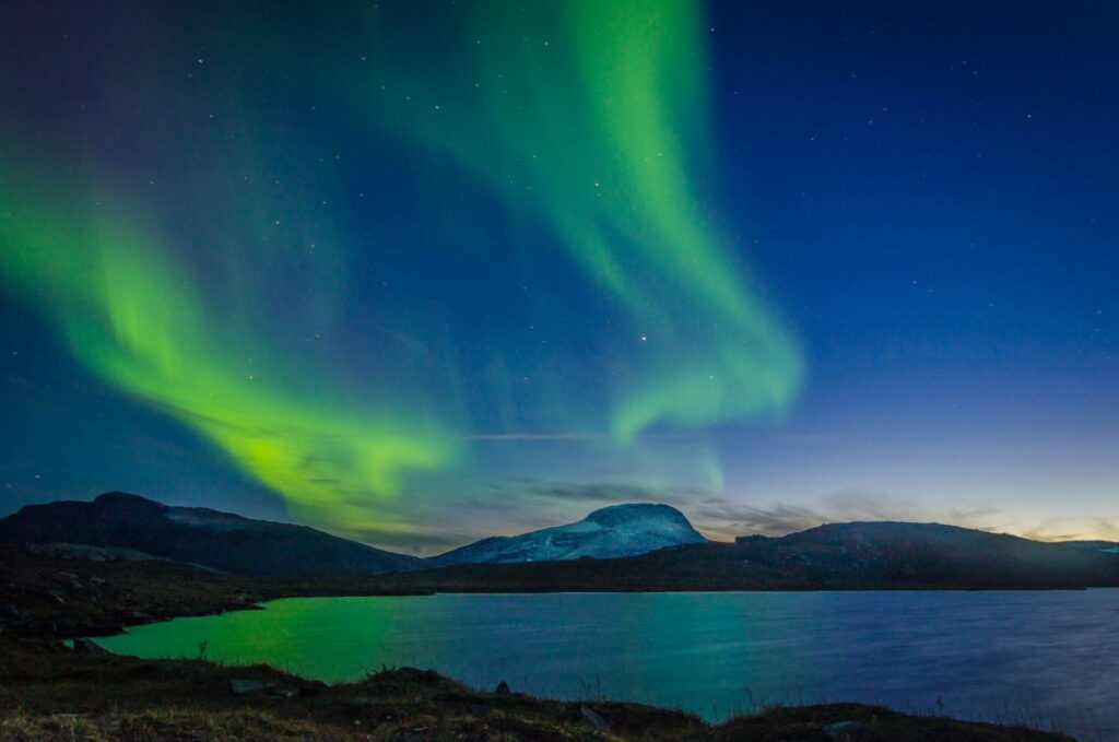 Aurora borealis over a tranquil lake with mountains in the background, likely in Sweden.