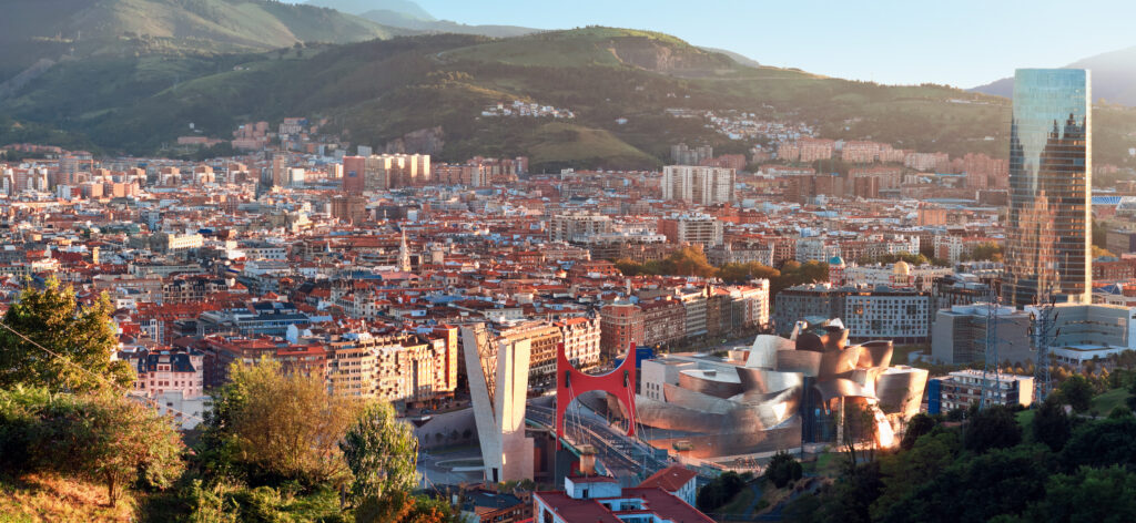 Panoramic view of Bilbao city with modern and traditional buildings, surrounded by green hills under a clear sky.