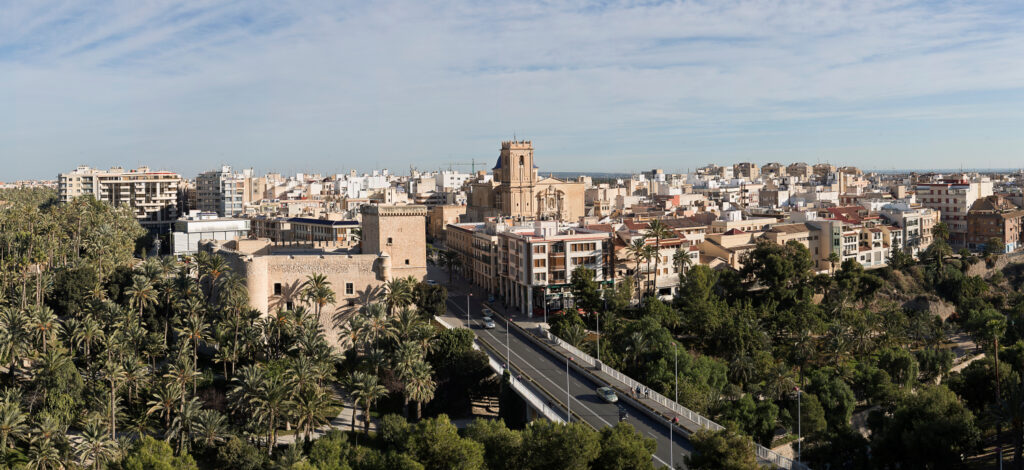 A panoramic view of Elche, Spain, features a cityscape with a central church, surrounded by greenery and trees.