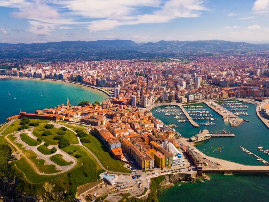 Aerial view of Gijon cityscape with buildings and marina, green park area on the left, and the sea surrounding the city.