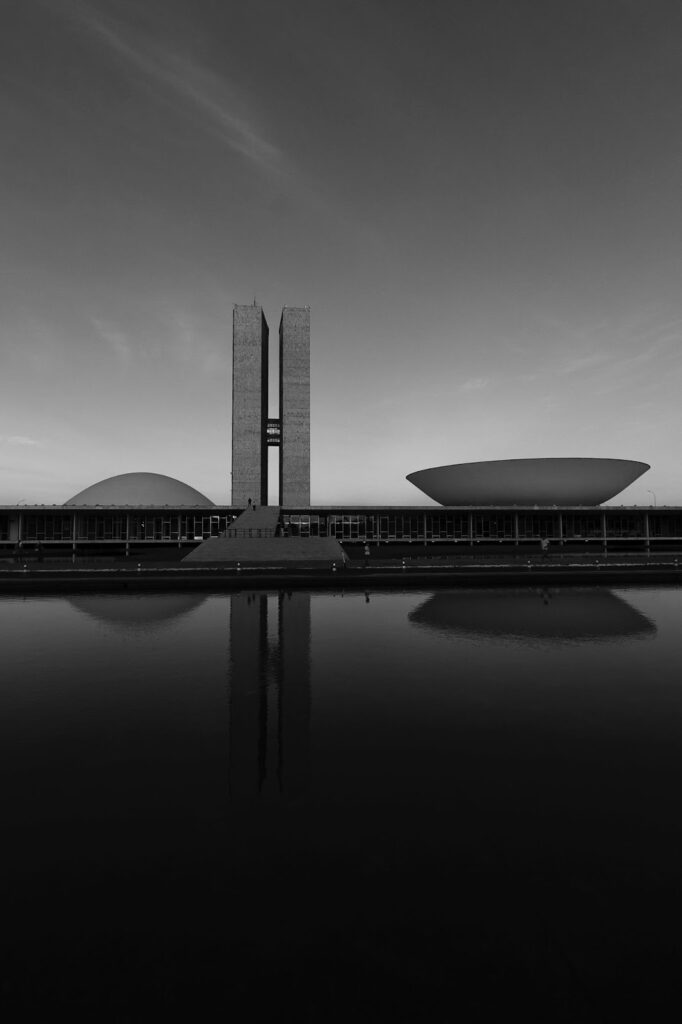 A black and white image shows the unique twin towers and dome of Brazil's National Congress in Brasília, reflected on a surface.