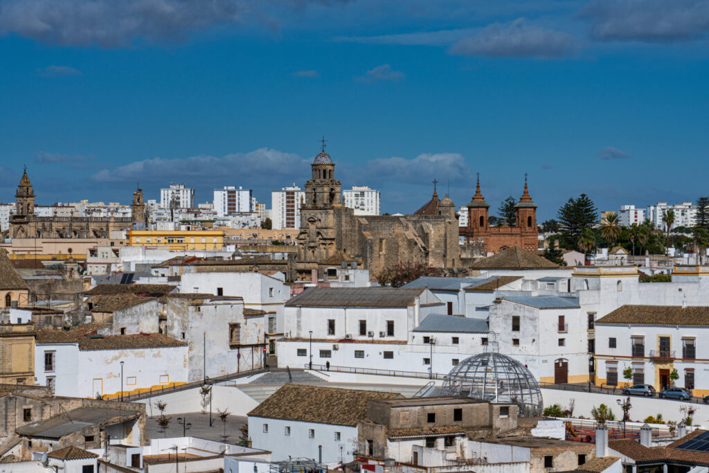 A panoramic view of Jerez de la Frontera features white Spanish buildings, churches with bell towers, under a blue, cloudy sky.