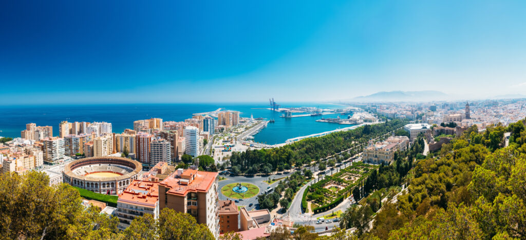 Panoramic view of Málaga city with coastline, buildings, and a bullring, under a clear blue sky.