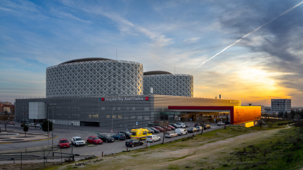 A unique, modern building with a geometric facade and orange section at sunset, likely in Móstoles, with a parking lot in front.