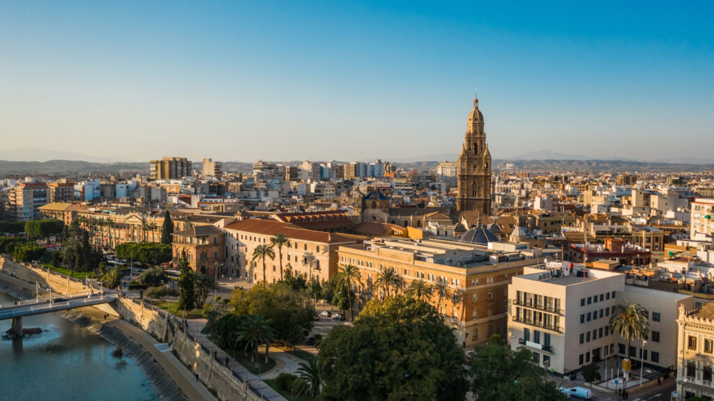 Aerial view of Murcia cityscape at sunset with a cathedral spire dominating the skyline and a river running alongside buildings.