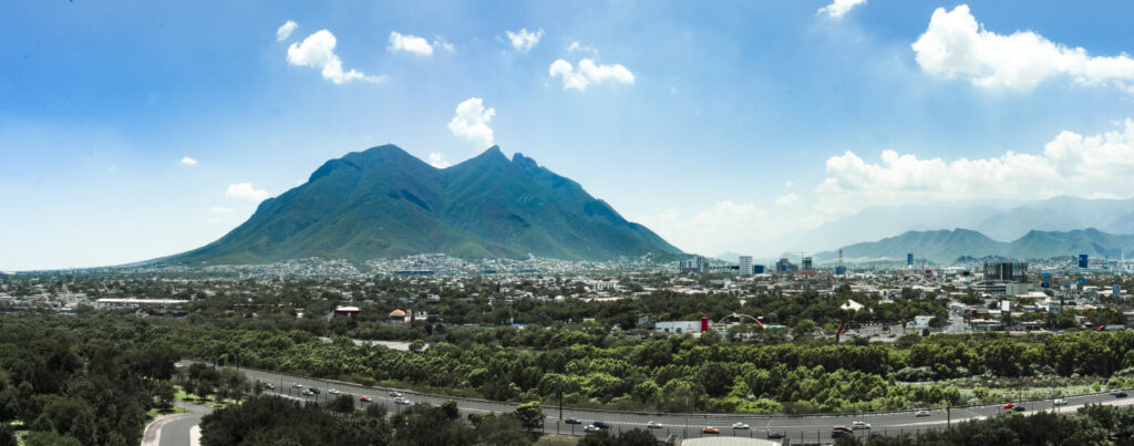 Panoramic view of a city with a large mountain in the background under a blue sky with scattered clouds, likely in Nuevo Leon, Mexico.