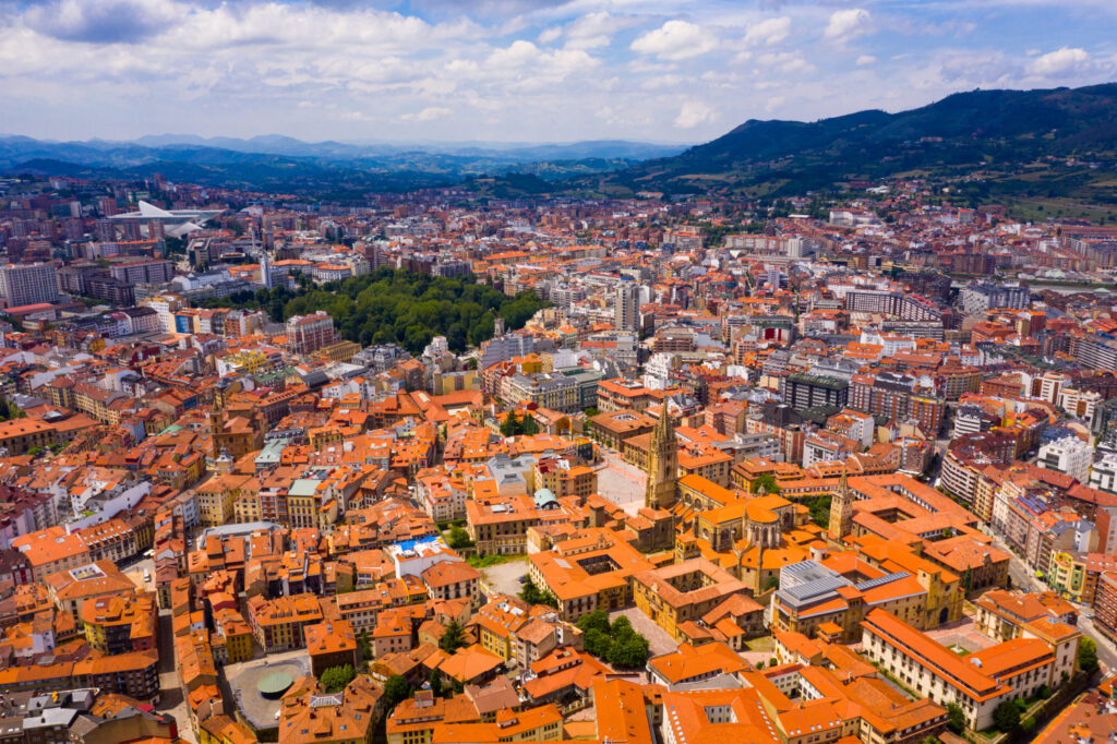 Aerial view of Oviedo cityscape with orange rooftops and surrounding green hills under a cloudy sky.