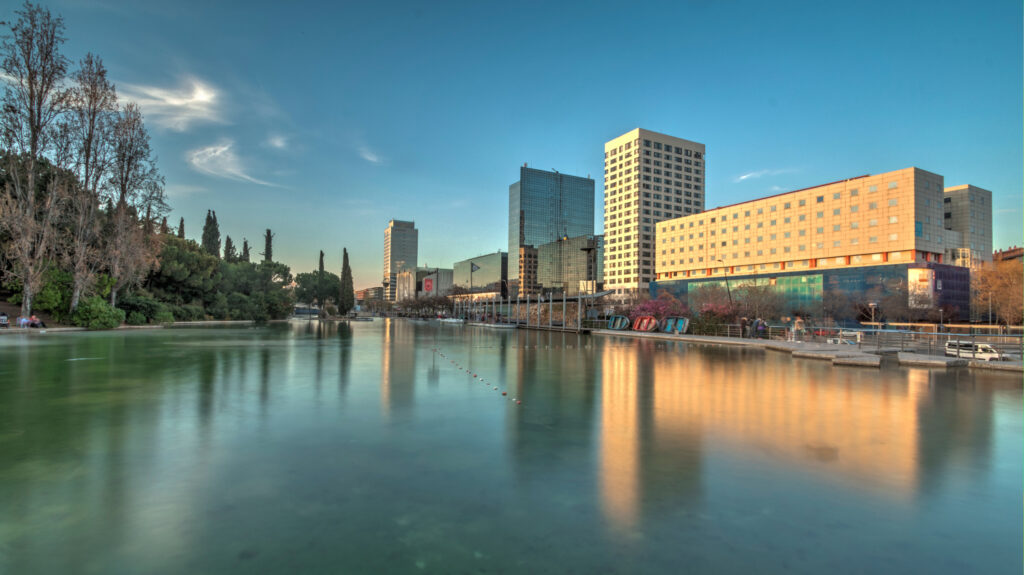 A cityscape of Sabadell with modern buildings reflected in a calm body of water, under a clear sky at dusk.