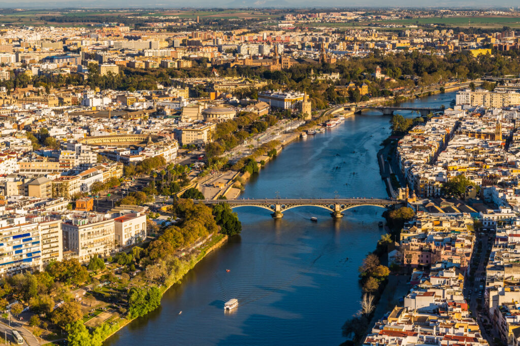 Aerial view of a river flowing through a city with bridges, buildings, and trees, likely in Sevilla.