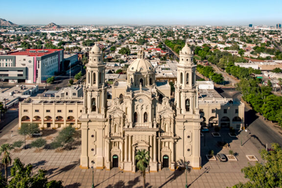 Aerial view of a grand cathedral with two bell towers in Sonora, Mexico, surrounded by a cityscape with buildings and trees under a clear blue sky.