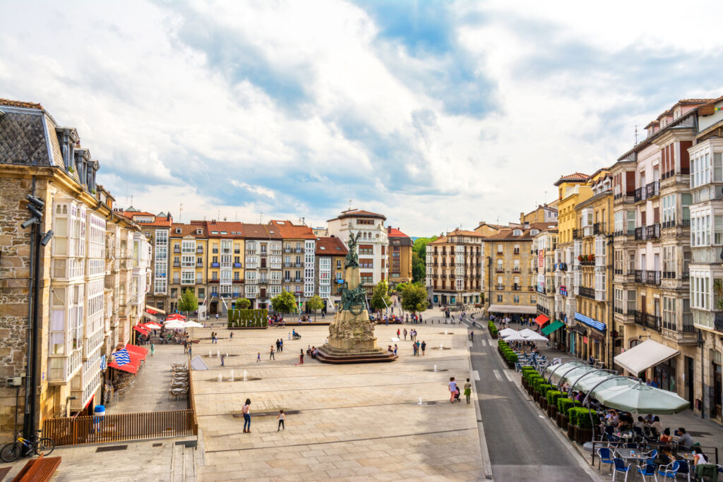 A vibrant town square in Vitoria-Gasteiz with people walking, buildings lining the perimeter, and a statue in the center under a partly cloudy sky.