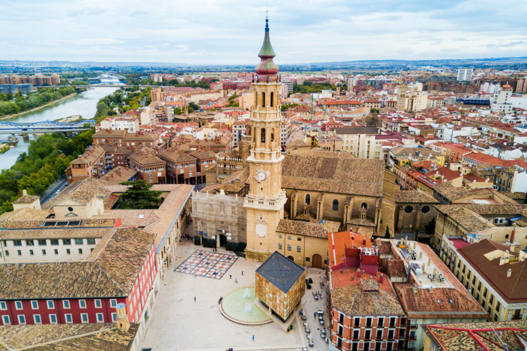 Aerial view of Zaragoza cityscape with historical buildings and a prominent church tower, near a river under a cloudy sky.