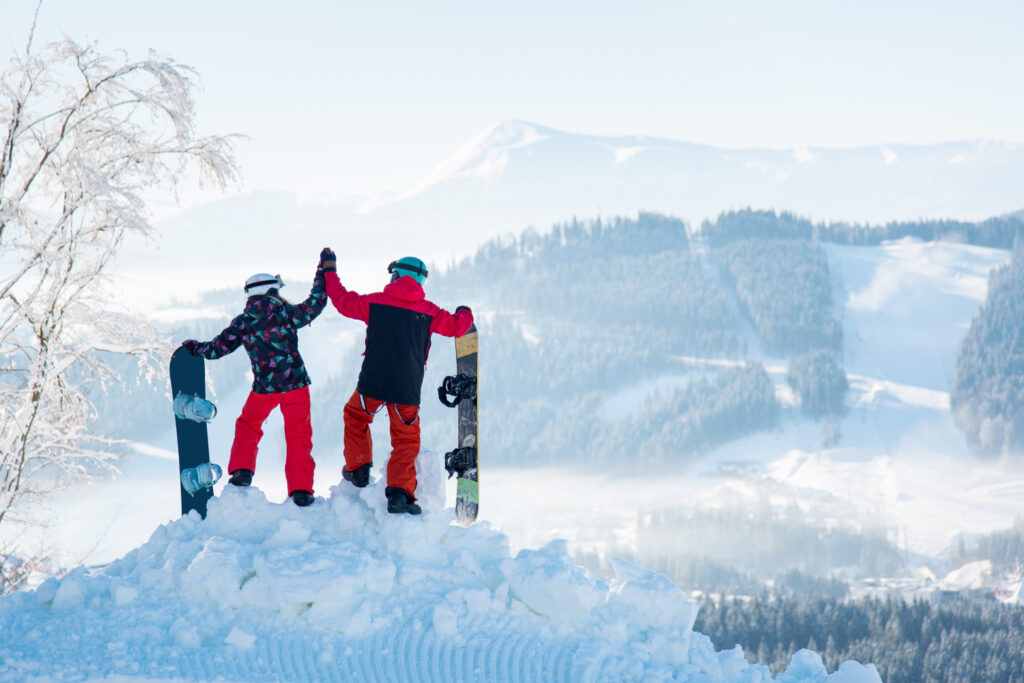 Couple snowboarders high fiving each other, standing on top of a snowy mountain, observing stunning winter view, resting after snowboarding at ski resort on a beautiful sunny winter day, Bukovel