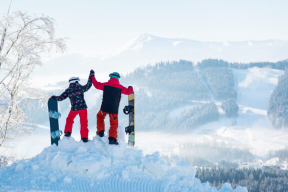 Three snowboarders stand on a snowy ridge at Bukovel Ski Resort, holding their boards and facing mountains.