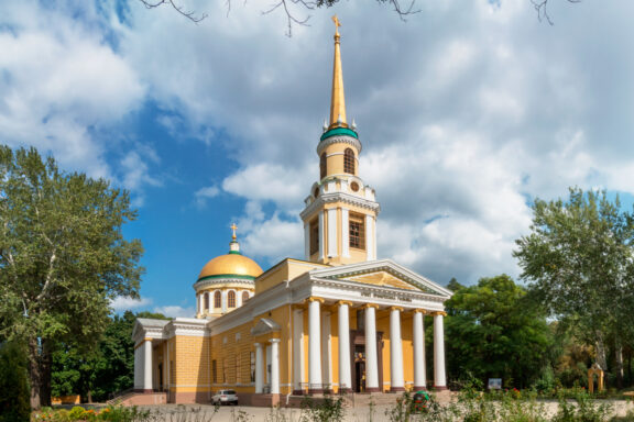 A cathedral in Dnipro boasts a tall spire, golden dome, classical architecture with columns, set against a blue sky.