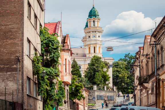 A Uzhhorod street view showcases a cathedral with a green dome, tower, buildings and trees under a partly cloudy sky.