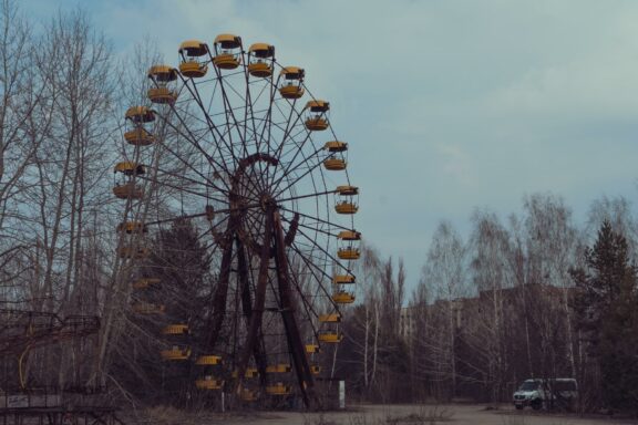 An old, abandoned Ferris wheel with yellow cabins in a desolate area with overgrown trees, under a cloudy sky, likely in Chernobyl.