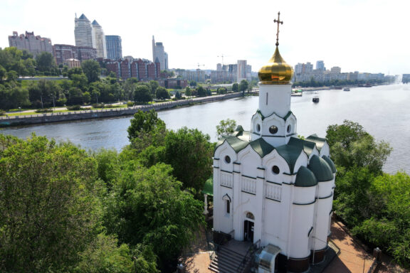 A white church with green domes and a golden cross, near a river with a city skyline, in Dnipro, Ukraine.