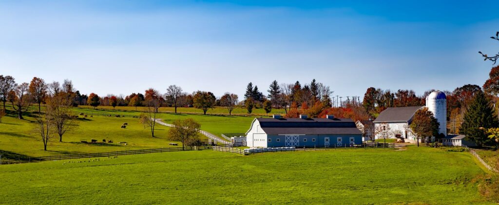 A panoramic view of a rural landscape in Connecticut, featuring a blue barn with white trim, a silo, and rolling green fields under a clear blue sky.