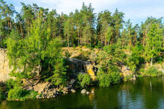 A scenic view of Korostyshiv Quarry with lush green trees surrounding a body of water under a clear sky.