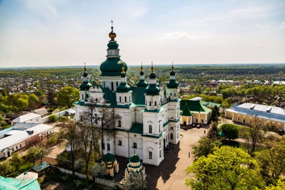 Aerial view of the Holy Trinity Cathedral with green domes, surrounded by trees, with a town in the background under a clear sky.