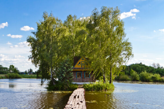 A small wooden house on an island in Solotvyn village, connected to the mainland by a footbridge and surrounded by greenery.