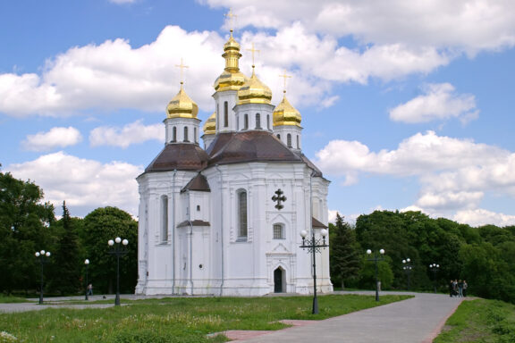 A white church with golden domes under a blue sky with scattered clouds, surrounded by greenery, possibly located in Kateryninska.