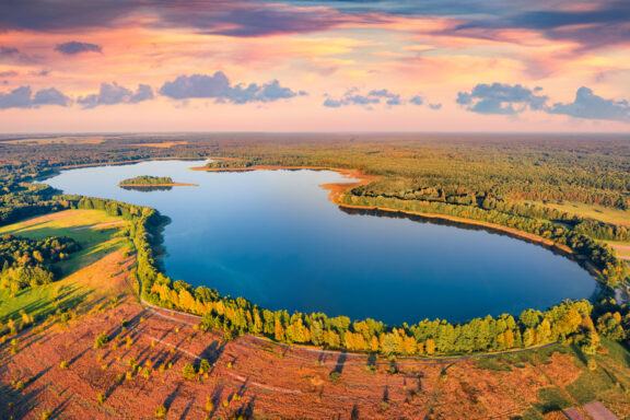 Aerial view of Krymne Lake at sunset with surrounding forest and fields, reflecting the sky's colors.