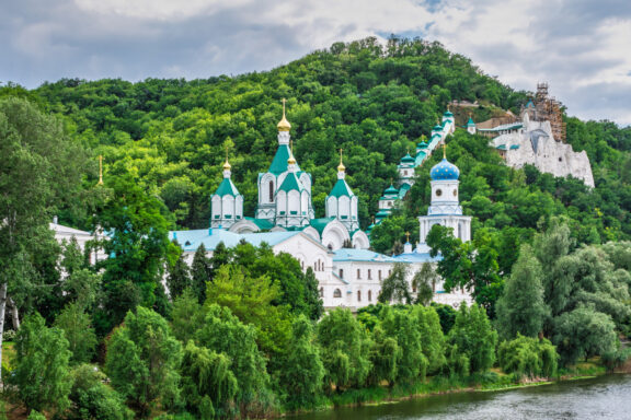 A picturesque view of the Svyatogorsk Lavra monastery, with its white buildings and blue domes, set amid greenery on a hill by a river.