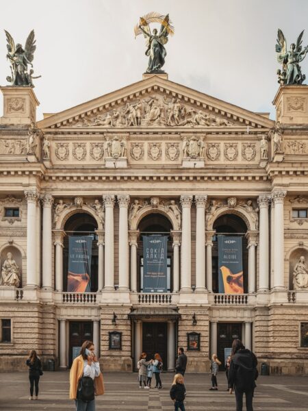 A photo showcases the grand neoclassical facade of Lviv National Opera House, with people strolling under a partly cloudy sky.