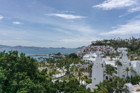 A coastal view of Manzanillo with white buildings nestled among green foliage, overlooking a calm blue sea under a partly cloudy sky.