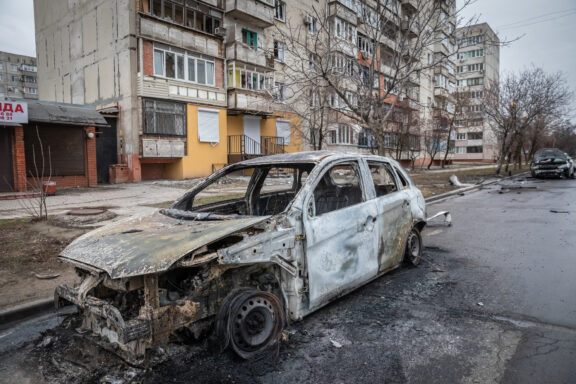 A scene of urban destruction, possibly from conflict or disaster, is suggested in Mariupol, Ukraine with a burnt-out car and damaged buildings.