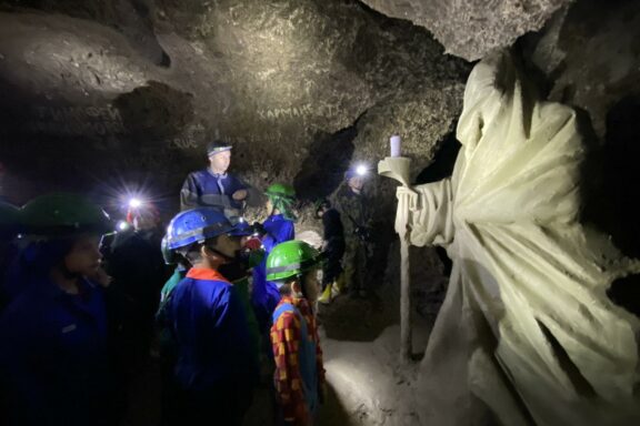 A group, with helmets and headlamps, explores the dimly lit, rocky Optimistic Cave, guided in the background.