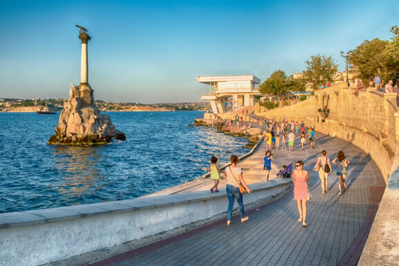 A seaside promenade in Sevastopol with people walking and enjoying the view, a monument on a rock in the sea, and a clear sky at dusk.