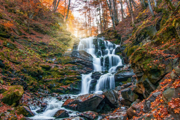 A cascading waterfall known as Shypit, surrounded by autumn-colored trees and rocks, with sunlight filtering through the foliage.