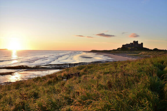 Sunset view of Bamburgh Castle by the sea with beach and grass in the foreground.