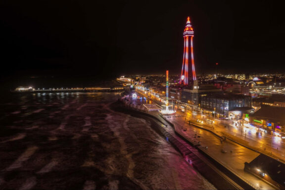 Nighttime view of Blackpool Tower illuminated in red and white lights, with the surrounding promenade and beach, located in Blackpool, England.