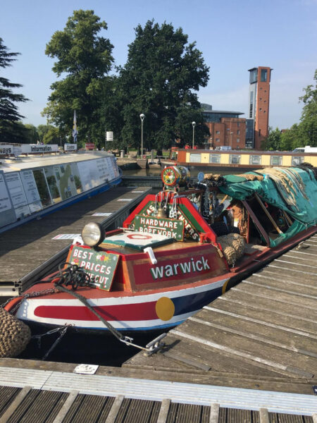 A narrowboat named Warwick moored at a dock with buildings and trees in the background on a sunny day.