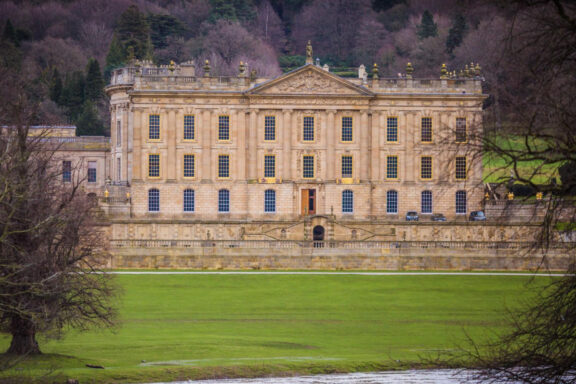 A stately mansion with classical architecture surrounded by a lush green lawn and trees, possibly located in the Chatsworth House gardens.